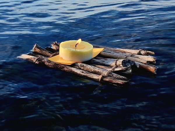 Eine brennende Kerze auf einem kleinen Floß aus Holzstöcken, das auf ruhigem Wasser treibt.
