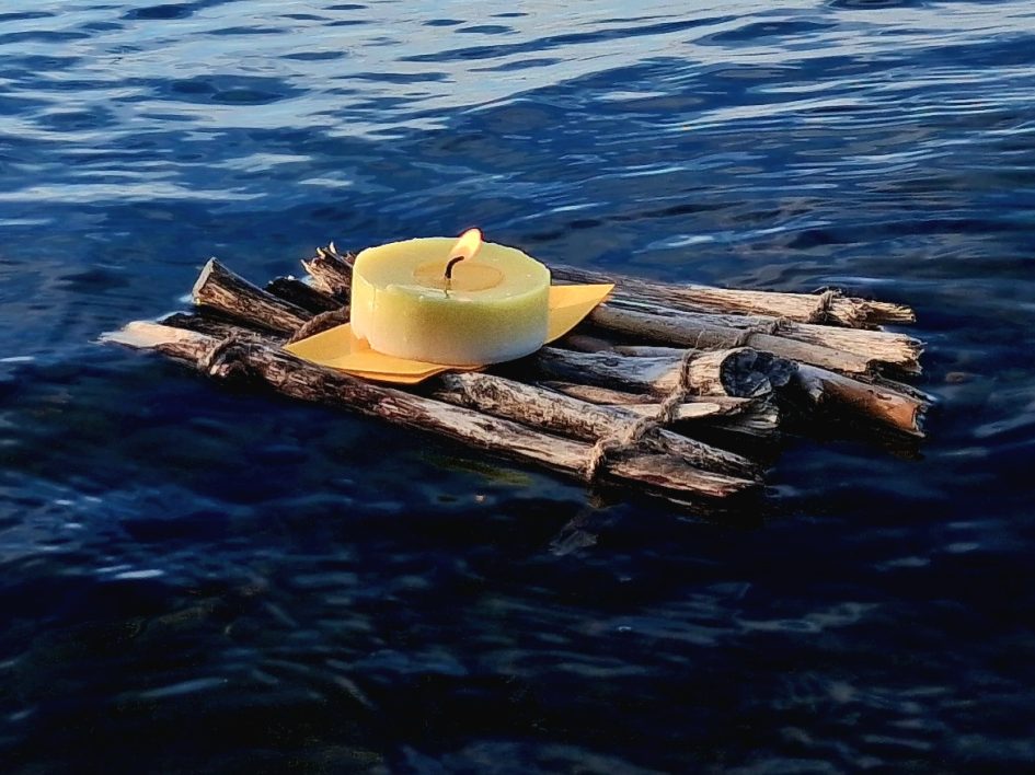 Eine brennende Kerze auf einem kleinen Floß aus Holzstöcken, das auf ruhigem Wasser treibt.
